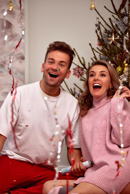 Una joven pareja enamorada se sienta en el suelo cerca del árbol de Navidad hace estallar una galleta de Año Nuevo y