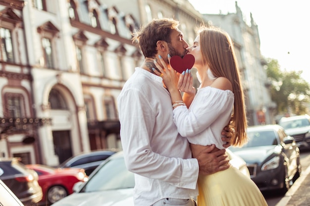Foto una joven pareja enamorada sostiene un corazón rojo muestran su amor y se besan en la ciudad concepto romántico