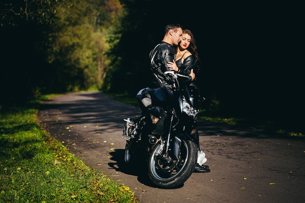 Joven pareja de enamorados abrazándose y besándose cerca de una motocicleta deportiva negra en el fondo de un camino forestal