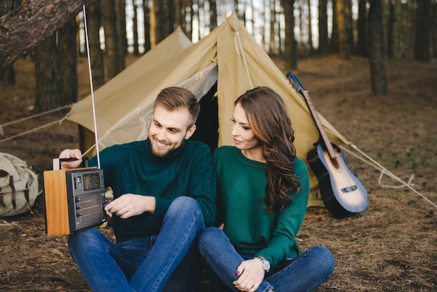 Joven pareja de enamorados acampando turistas sentados junto a un fuego contra una carpa en el bosque escuchando radio retro