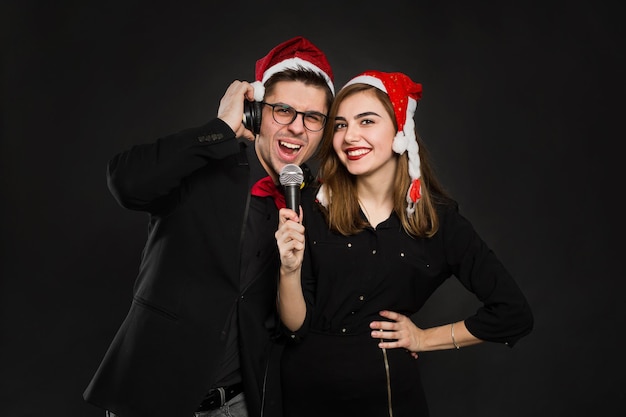 Joven pareja de enamorados está disfrutando de una fiesta de año nuevo, poniéndose gorro de Papá Noel y auriculares, sosteniendo un micrófono en sus manos.