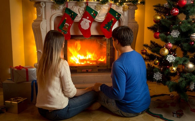 Joven pareja de enamorados sentados junto a la chimenea decorada o Navidad y mirando el fuego