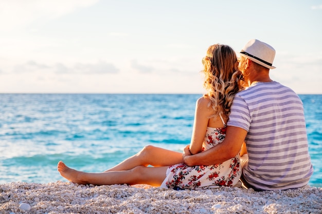 Joven pareja de enamorados sentados en la playa junto al mar disfrutando de una velada romántica y viendo la puesta de sol. La familia descansa en la playa en verano.