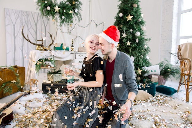 Joven pareja de enamorados sentados en el suelo cerca del árbol de Navidad lanzando confeti dorado