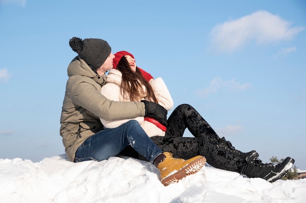 Joven pareja de enamorados se sienta en la nieve contra el cielo azul y se ríe.