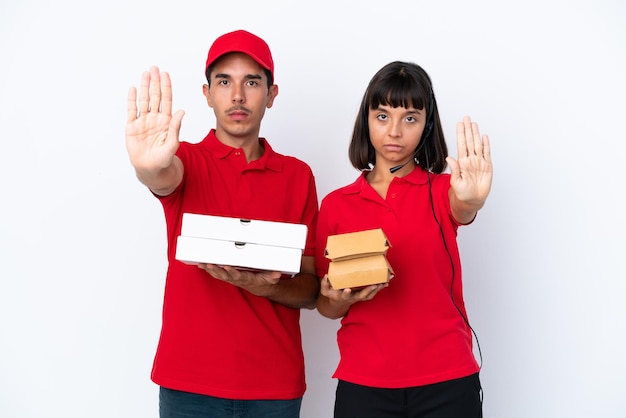 Joven pareja de entrega sosteniendo pizzas y hamburguesas aisladas en fondo blanco haciendo un gesto de parada negando una situación que piensa mal