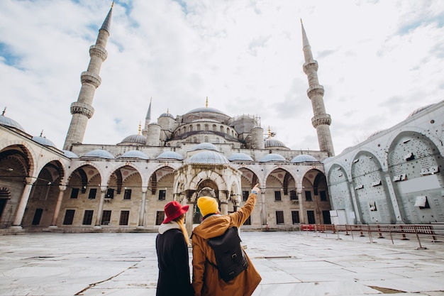 Una joven pareja europea camina en el patio de la Mezquita Azul en Estambul, Turquía. Viajero chico y chica con sombreros amarillos caminar en invierno Estambul. nublado día de otoño en Estambul.