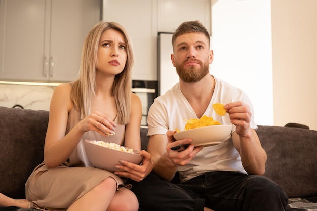 Joven pareja europea concentrada se sienta en el sofá y ve la televisión o una película. Chico y chica comiendo patatas fritas y palomitas de maíz. Ocio y descanso en casa. Concepto de disfrutar el tiempo juntos. Interior del apartamento estudio