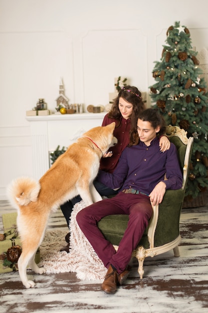 Joven pareja feliz abrazando al adorable perro akita inu mientras se sienta en un elegante sillón retro para las vacaciones de Navidad en casa, el árbol de Navidad y la chimenea, acogedoras decoraciones para el hogar