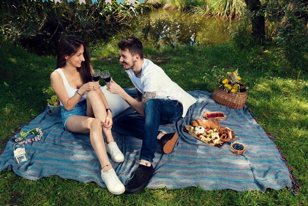 Foto una joven pareja feliz bebiendo vino rojo en un picnic en el parque de la ciudad sentada en una manta enfoque selectivo