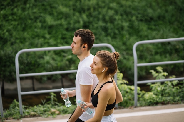 Joven pareja feliz corriendo en el parque de la ciudad con una botella de agua en las manos, deportes conjuntos, alegría, estilo de vida saludable del deporte de la ciudad, fitness juntos en las noches de verano, corredores