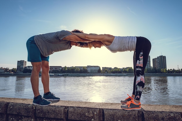 Joven pareja feliz entrenando al aire libre junto al río, estirándose juntos en la pared al atardecer.