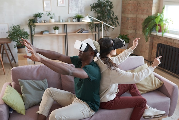 Joven pareja feliz en gafas sentado en el sofá y gesticulando mientras juega un juego de realidad virtual en casa