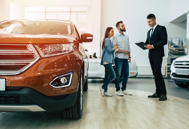 Joven pareja feliz moderna comprando un auto nuevo en el concesionario de la ciudad y hablando con el vendedor