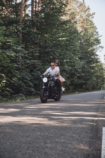 Una joven pareja feliz monta una motocicleta en una carretera asfaltada en el bosque libertad y velocidad