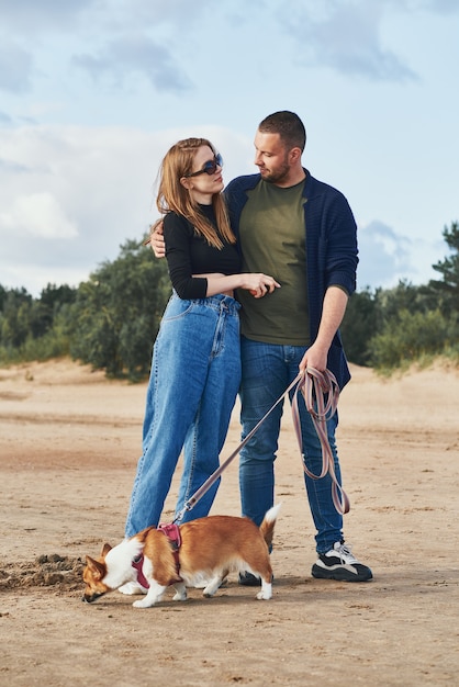 Joven pareja feliz y perro parados en la playa contra pinos y arena. Hombre guapo y mujer hermosa