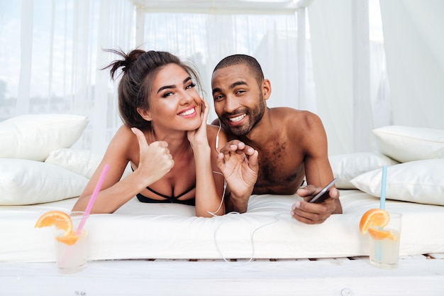 Joven pareja feliz sonriente tendido en la cama de la playa y mostrando signo bien