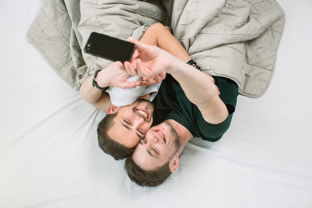 Joven pareja gay acostado debajo de la manta en la cama en la sala de luz un selfie haciendo en el teléfono inteligente. Concepto de pareja gay, homosexual