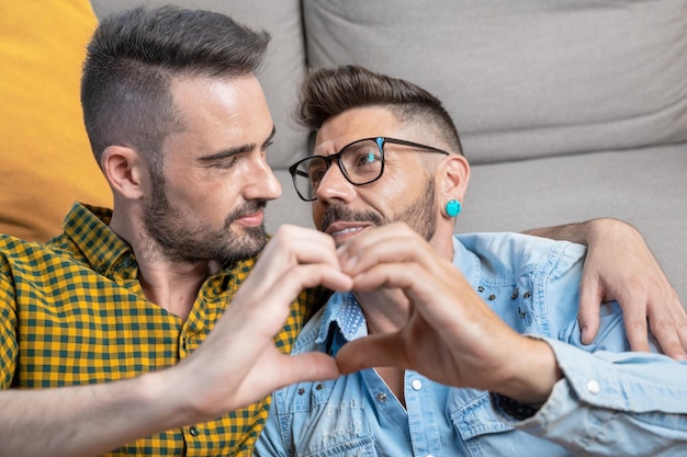 Joven pareja gay sonriendo confiada haciendo el símbolo del corazón con las manos en casa