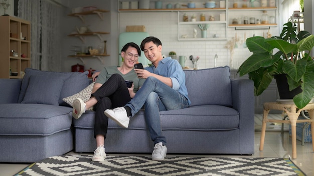 Joven pareja gay sonriente usando un teléfono inteligente y relajándose en la sala de estar en casa
