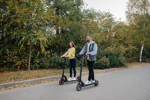 Una joven pareja hermosa paseos en patinetes eléctricos en el parque en un cálido día de otoño