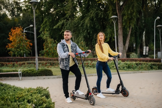 Una joven pareja hermosa paseos en patinetes eléctricos en el parque en un cálido día de otoño