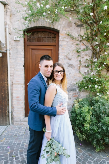 Joven pareja hermosa en ropa azul sonriendo y de pie en el fondo del antiguo edificio, el hombre abraza a la mujer. Sirmione, Italia