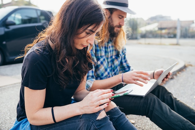 Joven pareja hermosa sentada al aire libre con computadora y teléfono inteligente