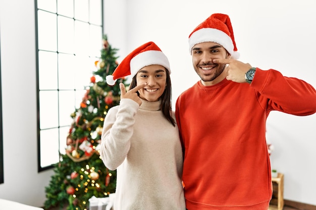 Joven pareja hispana de pie junto al árbol de navidad apuntando con el dedo de la mano a la cara y la nariz sonriendo alegre concepto de belleza