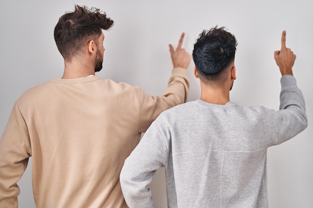 Joven pareja homosexual de pie sobre fondo blanco posando hacia atrás apuntando hacia adelante con la mano del dedo