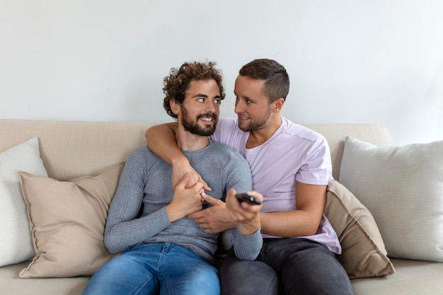 Joven pareja homosexual relajándose en el sofá de la sala de estar abrazándose y disfrutando mientras ve la televisión
