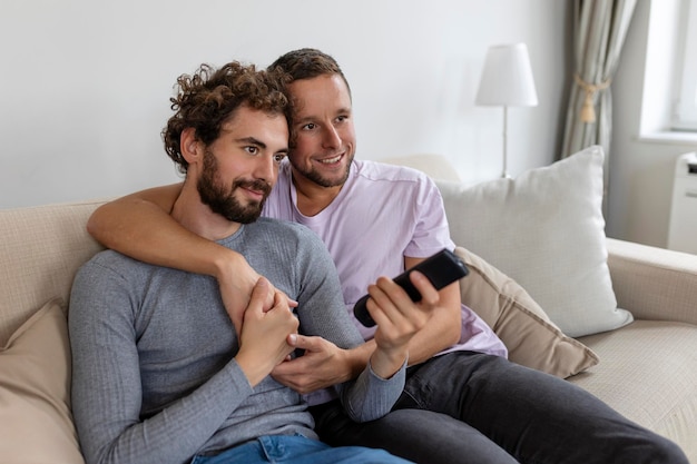 Joven pareja homosexual relajándose en el sofá de la sala de estar abrazándose y disfrutando mientras ve la televisión