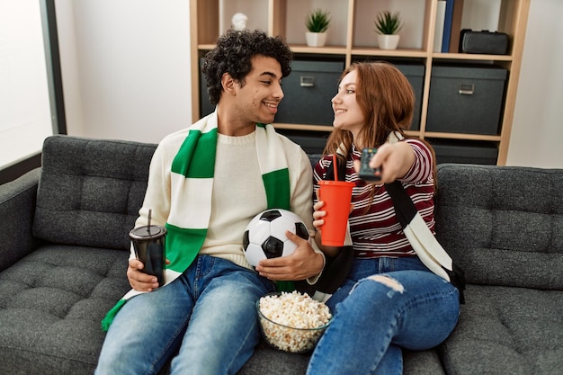 Joven pareja hooligan sonriendo feliz apoyando al equipo de fútbol sentado en el sofá en casa