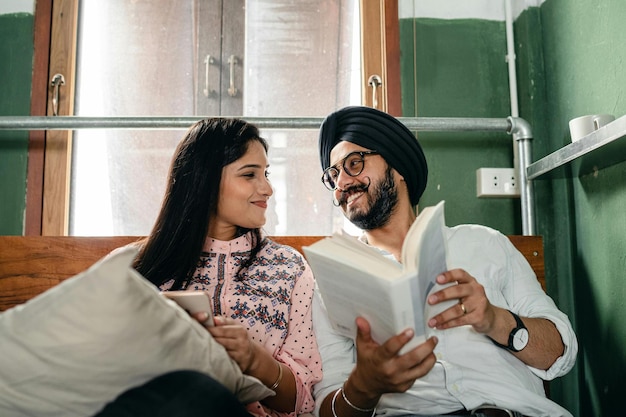 Foto una joven pareja india amorosa con ropa casual y turbante con un libro y un teléfono móvil descansando en una cama cómoda