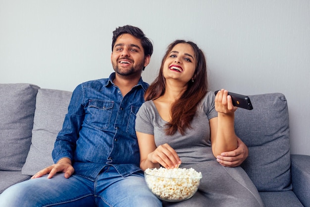 Joven pareja india viendo una película de comedia con control remoto en la mano en la sala de estar con palomitas de maíz