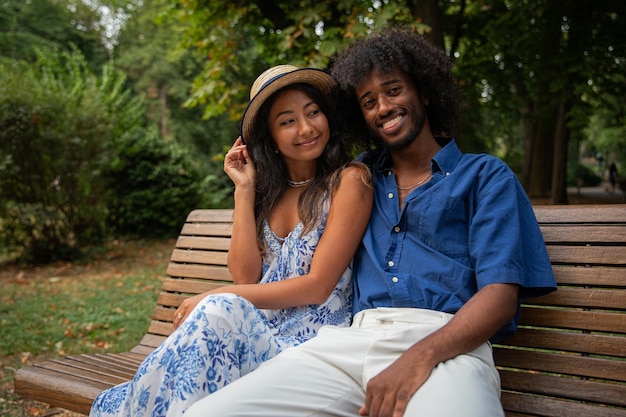 Una joven pareja interracial, él es africano, ella es asiática, están sentados en un banco en un parque público y sonriendo.