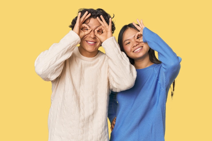 Foto una joven pareja interracial en el estudio mostrando el signo de "okay" sobre los ojos.