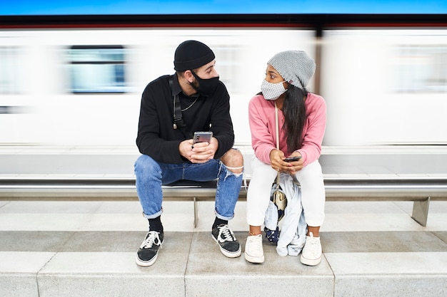 Una joven pareja interracial con máscaras y gorros de lana se sienta en la plataforma del metro hablando