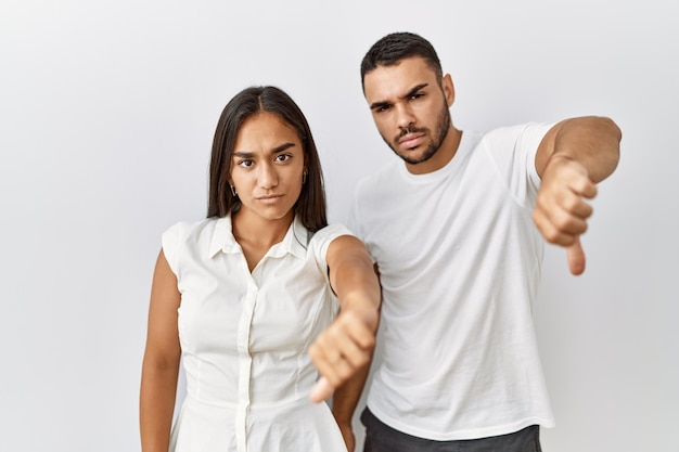 Joven pareja interracial de pie juntos enamorados sobre un fondo aislado que se ve infeliz y enojado mostrando rechazo y negativo con el pulgar hacia abajo gesto mala expresión