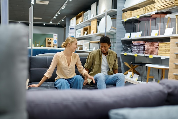 Joven pareja interracial sentada eligiendo el mejor sofá para el hogar en la tienda. Compra de muebles