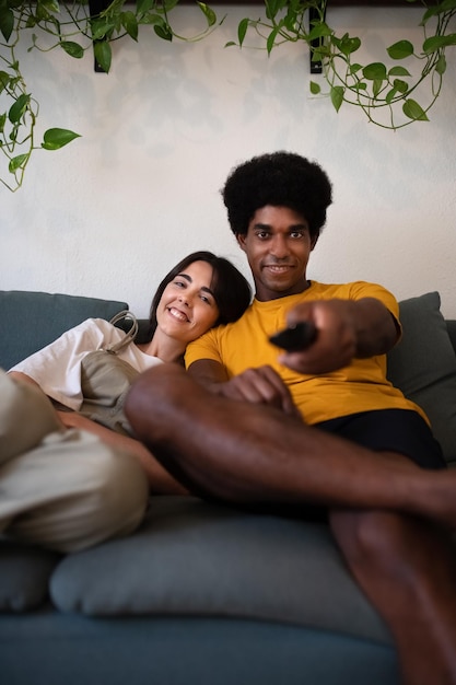 Joven pareja interracial ve la televisión en casa. Hombre afroamericano cambia de canal con control remoto. Imagen vertical. Concepto de estilo de vida.