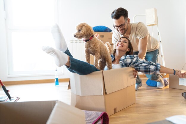 Una joven pareja juguetona en su nuevo apartamento jugando con su lindo perro en cajas de embalaje