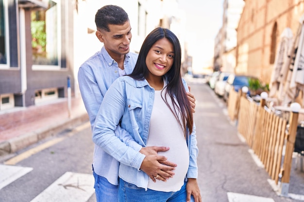 Joven pareja latina esperando bebé abrazándose unos a otros de pie en la calle