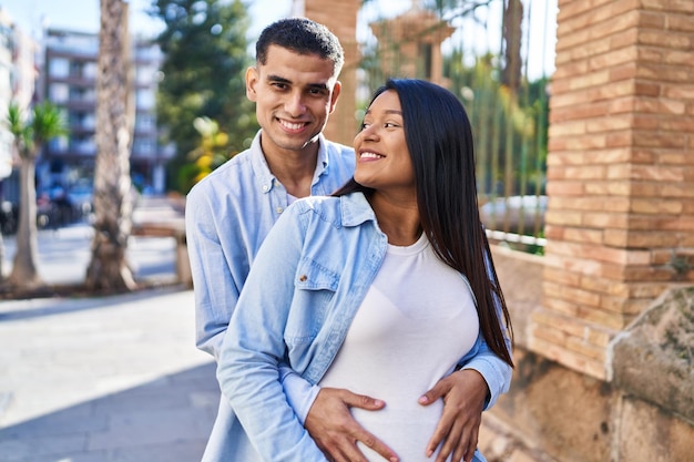 Joven pareja latina esperando bebé abrazándose unos a otros de pie en la calle