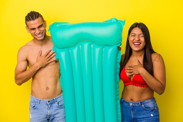 Joven pareja latina sosteniendo colchón de aire aislado sobre fondo amarillo se ríe a carcajadas manteniendo la mano en el pecho.