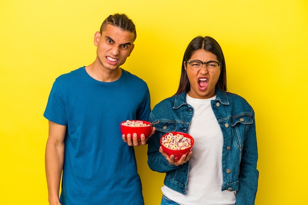 Joven pareja latina sosteniendo un tazón de cereales aislado en la pared amarilla gritando muy enojado y agresivo.