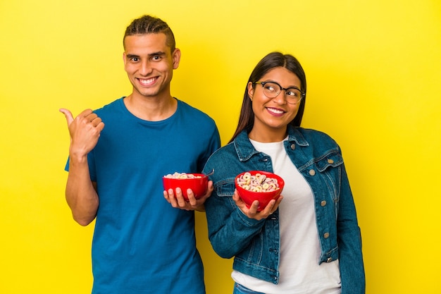 Joven pareja latina sosteniendo un tazón de cereales aislado en puntos de fondo amarillo con el dedo pulgar de distancia, riendo y despreocupado.