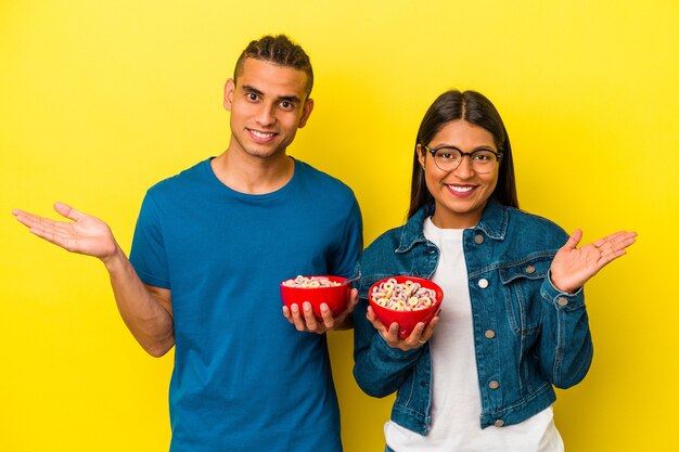 Joven pareja latina sosteniendo un tazón de cereales aislado sobre fondo amarillo que muestra un espacio de copia en una palma y sosteniendo otra mano en la cintura.