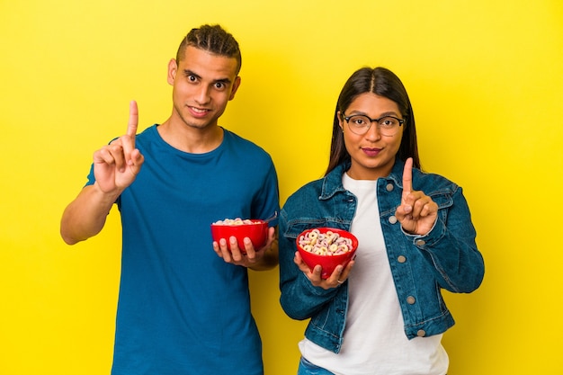 Joven pareja latina sosteniendo un tazón de cereales aislado sobre fondo amarillo que muestra el número uno con el dedo.