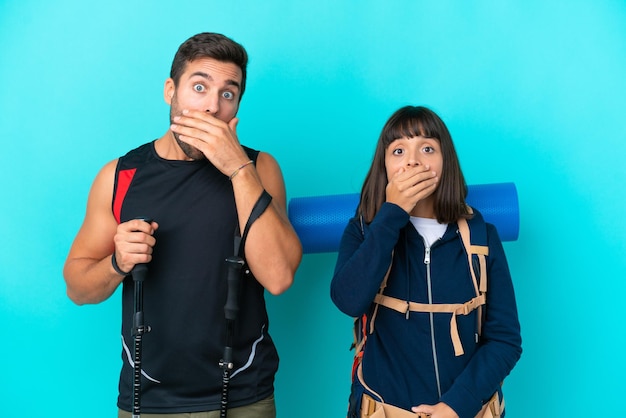 Joven pareja de montañeros con una mochila grande aislada de fondo azul cubriendo la boca con las manos por decir algo inapropiado